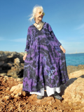 Woman in a purple tie-dye dress standing on rocks by the ocean.