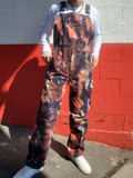 Person wearing a tie-dye overall in front of a red-white brick wall.