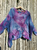 A loose, open-knit long-sleeve top with a blue, purple, and pink tie-dye pattern, hanging on a wooden hanger against a weathered wooden fence.