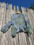 A hand-knit long-sleeve sweater with a blue, green, and beige tie-dye pattern, hanging on a wooden fence against a clear blue sky.