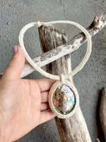 Hand holding a macrame necklace with a pendant on a piece of driftwood.