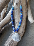 Necklace with blue beads and a shell pendant on a piece of driftwood.