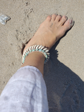 Foot wearing a shell bracelet on a sandy surface