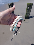 Hand holding an anklet with shell and amazonite beads on a beach.