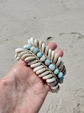 A hand holdinga beach anklet made of white shell beads and light blue beads, photographed outdoors on a sandy beach background.