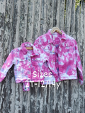 2 different size kids tie dye denim jackets displayed on our wooden fence. They are tie dyed in Barbie pink & light wisteria colors. The jackets feature all the usual jacket details: collar, metal buttons, cuffs, chest pockets with button flaps, and side pockets