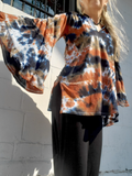 Person wearing a tie-dye shirt with bell sleeves, in orange, black, and white color combo against a light-colored wall.