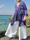 Person wearing a purple tie-dye shirt and white pants standing on rocks by the ocean.
