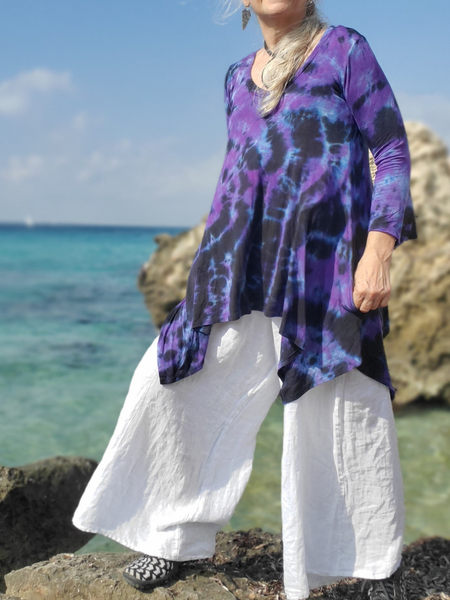 Person wearing a purple tie-dye shirt and white pants standing on rocks by the ocean.