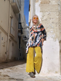 Person wearing a colorful tie-dye shirt and yellow pants standing in a narrow street.