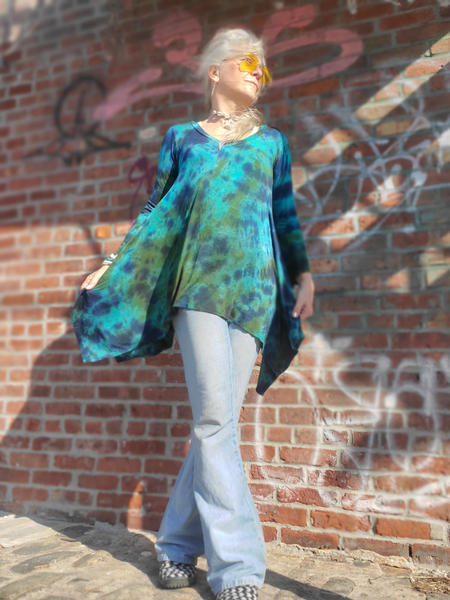Person wearing a blue tie-dye shirt and light blue jeans standing against a brick wall with graffiti.