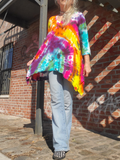 Person wearing a colorful tie-dye shirt and jeans standing against a brick wall.