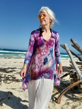 Woman in a colorful tie-dye shirt and white pants standing on a beach with ocean and clear sky in the background.