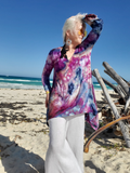 Woman in a tie-dye shirt and white pants standing on a beach with ocean and clear sky in the background.