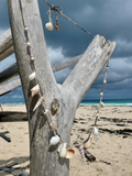Decorative shell garland hanging from a wooden post on a beach with ocean and sky in the background.