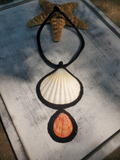 A handmade seashell necklace featuring a large white scallop shell and a smaller orange-pink clam shell, both bordered in black rope, with a black braided loop. It is displayed on a weathered gray wooden surface next to a brown starfish.