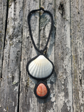 A handmade crocheted pendant necklace featuring two seashells, one larger white scallop shell and one smaller orange-brown scallop shell, both framed in black macrame and hanging against a weathered gray wooden plank background.