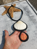 A handmade seashell necklace with a black crocheted border, featuring a large white shell and a smaller orange shell, shown resting on a gray surface with a hand holding one of the shells and a starfish nearby.