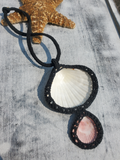 A handmade macrame necklace featuring a large white seashell and a smaller pink seashell, both framed in black woven cord, displayed on a weathered gray wooden surface next to a starfish.