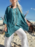 Woman in teal top and white pants standing on a beach with blue sky and ocean in the background.
