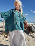 Woman standing on a beach wearing a teal top and white pants with a necklace.