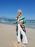 Woman in a colorful cardigan and white pants standing on a beach with blue sky and ocean.