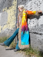 Person wearing a colorful tie-dye coat with fur trim against a textured wall.