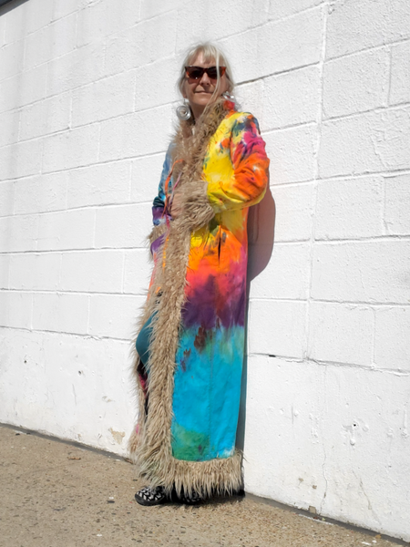 Person wearing a tie-dye dress with a long, fringed scarf against a white wall.