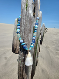 Driftwood with blue and green beads and a white shell on a sandy beach.