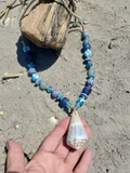 Necklace with blue and green beads and a shell pendant held by a hand on sandy ground.