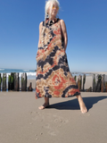A woman stands on a sandy beach wearing a sleeveless maxi dress with a tie-dye pattern in beige, orange, black, and brown. The background shows the ocean, wooden pilings, and a clear blue sky.