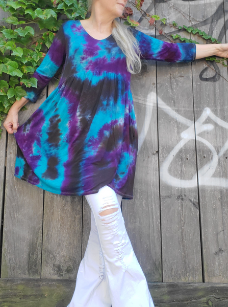I'm modeling a ¾ sleeve sleeve babydoll dress with a scoop neck, side pockets, mini lenght. I have a pair of white jeans under. The originally heather grey dress is had dyed in black-turquoise-purple colors. I'm standing front of a grey fence, holding out the skirt part, lifting up my other arm.