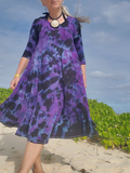 Woman wearing a purple and black patterned dress standing on a beach with greenery and blue sky in the background.