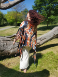 A woman outdoors in a grassy park wearing a colorful tie-dye fringe poncho and white skirt, with trees and fallen logs in the background.
