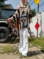 Person wearing a tie-dye poncho with fringe, white pants, and sandals on a street.
