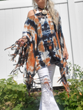 A woman wearing a bohemian-style tie-dye poncho top with fringe, paired with white distressed jeans. The top features a black, orange, and white tie-dye pattern with long brown fringe.