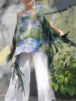 Person wearing a colorful one-shoulder top with fringes and white pants, standing against a textured wall with greenery.