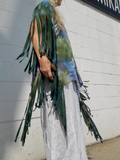 Person wearing a colorful top with fringes and white pants against a gray wall.