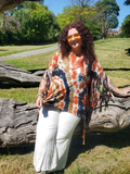 A woman with curly red hair and tattoos wearing a tie-dye fringed wrap top and white pants, leaning on a large fallen tree trunk in a grassy park.