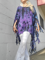 Person wearing a purple tie-dye poncho with fringe against a gray wall.