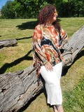 A woman with curly red hair and tattoos wearing a tie-dye fringed wrap top and white pants, leaning on a large fallen tree trunk in a grassy park.