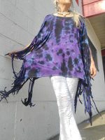 Person wearing a tie-dye poncho with fringe details against a gray wall.