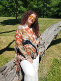 A woman with curly red hair and tattoos wearing a tie-dye fringed wrap top and white pants, leaning on a large fallen tree trunk in a grassy park.