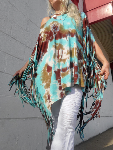 Person wearing a tie-dye poncho with fringe in front of a gray wall.