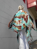 Person wearing a colorful tie-dye poncho top with fringe, standing against a gray wall.
