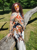 A woman with curly red hair and tattoos wearing a tie-dye fringed wrap top and white pants, leaning on a large fallen tree trunk in a grassy park.