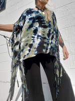 Person wearing a tie-dye poncho with fringe details against a white wall.