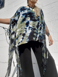 Person wearing a tie-dye poncho with fringe details against a white wall.