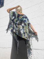 Person wearing a tie-dye poncho with fringe on a white brick wall background, a back shot