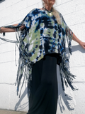 Person wearing a  sage-black tie-dye poncho with fringe against a white wall